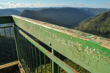 Fototapeta premium Carved markings on the railing of Gelain Lookout, with a scenic view of forested hills and a deep valley in Flores da Cunha, Brazil