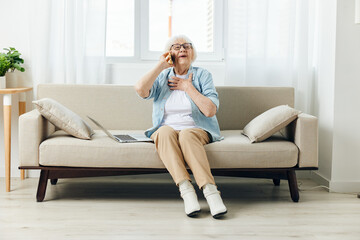 Obraz premium a joyful elderly lady is sitting on a cozy sofa in a bright interior and smiling talking on the phone with her hand on her chest