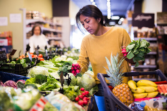 Portrait of pensive latin american woman buying fresh organic vegetables in supermarket, choosing red radish