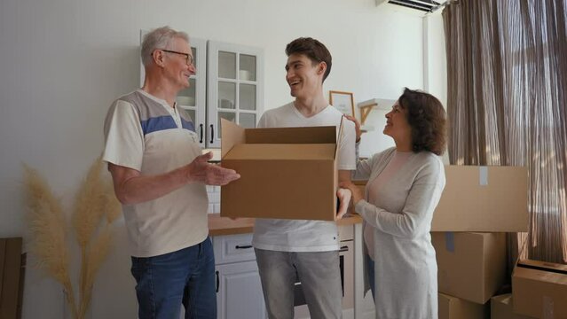 Young man holds cardboard box talking with elderly parents in kitchen of new apartment. Happy senior couple waves goodbye to son walking away with box
