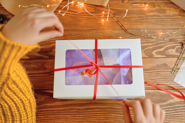 Girl in orange sweater pulls ribbon on gift box. First-person view of woman's hands opening Christmas surprise. Wooden table with garland. New Year's holiday mood.