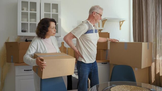 Elderly man carries heavy cardboard boxes complaining of pain in back to woman. Senior couple puts things in order in kitchen after relocating to new apartment