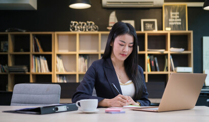 Obraz premium Professional businesswoman using laptop computer and taking notes on notebook at her office desk.