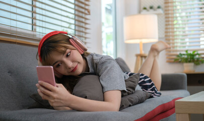 Happy young woman relaxing on couch in living room, wear bluetooth headset and using smartphone.