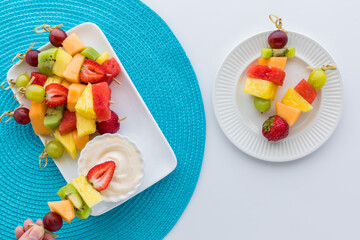 Fresh fruit kebabs on a platter with a hand dipping one into a yogurt dip.