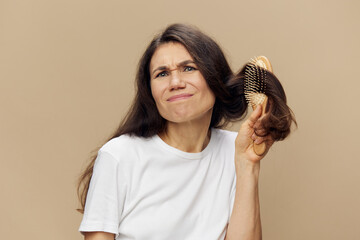 A sad, upset woman stands with tangled hair in a comb and with an emotionally depressed face trying to untangle her hair standing on a beige background