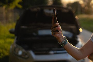 side view of hand of unknown caucasian woman holding mobile phone against opened car hood broken...