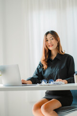 Smiling happy Asian woman relaxing using the technology of a laptop computer while sitting in a chair at a desk. creative girl working at home The concept of working and studying online learning