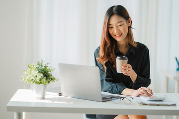 Smiling happy Asian woman relaxing using the technology of a laptop computer while sitting in a...