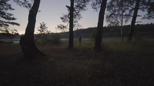 Distant shot of a woman taking photos with her cell phone in a forest at sunset.