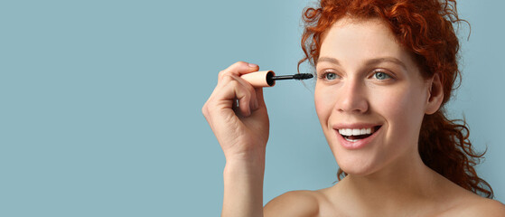 Beautiful redhead woman applying mascara against blue background with space for text