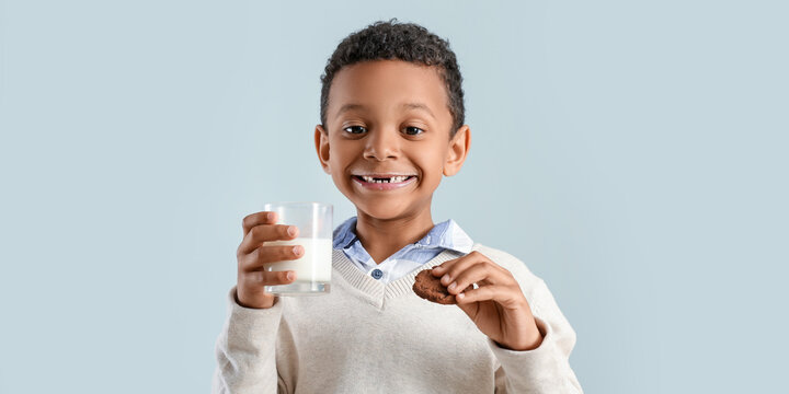 Cute African-American boy drinking milk and eating tasty cookie on light background