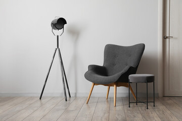 Comfortable armchair with stool and lamp near light wall in room