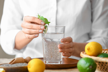 Woman adding mint leaves into glass with water and chia seeds on table