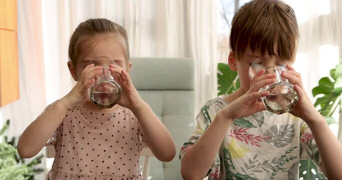 Brother and sister sit at kitchen table drinking water from glasses. Children quench thirst before going to school in morning closeup slow motion