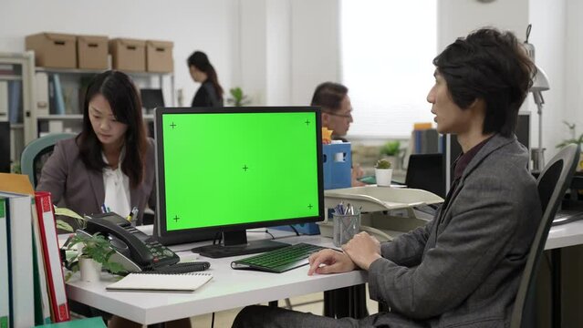 Asian male manager wearing suit talking with hand gestures while having web conference via video chat on computer with chroma key green mockup in the company