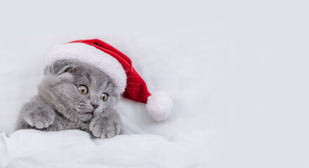 Cozy kitten wearing red santa's hat lying under a white blanket on a bed and looks away on empty...