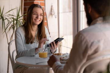 young joyful couple using cellphones and laughing while having breakfast in kitchen at home, bearded man and woman eat healthy food and drink juice