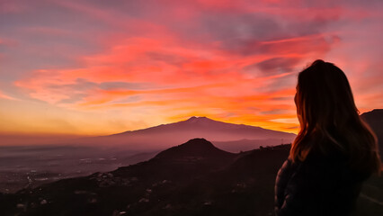 Fototapeta premium Tourist woman watching beautiful sunset behind volcano Mount Etna near Castelmola, Taormina, Sicily, Italy, Europe, EU. Clouds with vibrant red orange colors. Silhouette of person during twilight