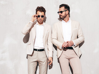 Portrait of two handsome confident stylish hipster lambersexual models. Sexy modern men dressed in white elegant suit. Fashion male posing in studio near grey wall. In sunglasses