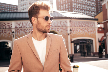 Handsome confident stylish hipster lambersexual model.Sexy modern man dressed in elegant beige suit. Fashion male posing in the street background in Europe city at sunset. In sunglasses