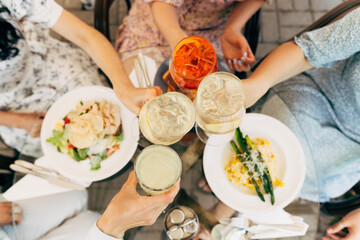 Overhead view of friends having brunch, toasting, saying cheers holding tropical aperol spritz cocktails. Food and beverage celebration concept