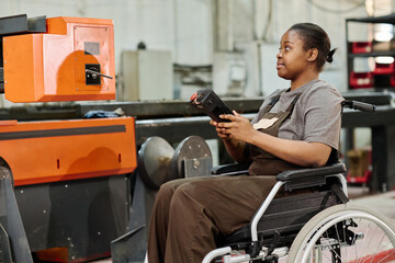 African female operator in wheelchair controlling the work of machine with console, she working in industrial factory