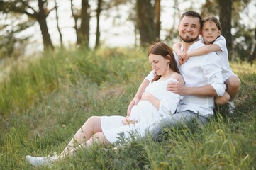 Fototapeta premium Beautiful pregnant brunette mom and dad and dauther in white clothes look at each other in nature.