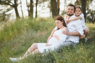 Beautiful pregnant brunette mom and dad and dauther in white clothes look at each other in nature.