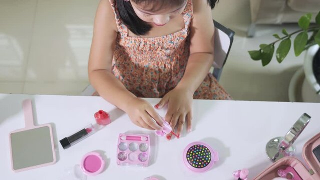 Asian adorable funny little girl making makeup dips brush into bottle to paints nails polish red nail varnish herself, Learning activity to be woman, happy kid is beautiful make up with cosmetics toy
