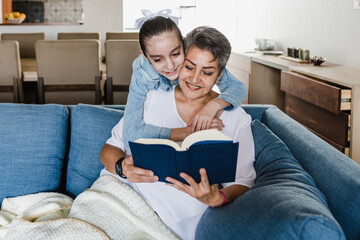hispanic Grandmother and granddaughter reading a book on sofa at home in Mexico Latin America