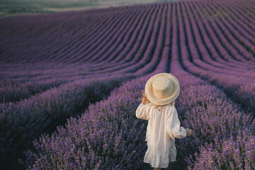 Happy girl in straw hat and white dress enjoying flower fragrance in lavender field.