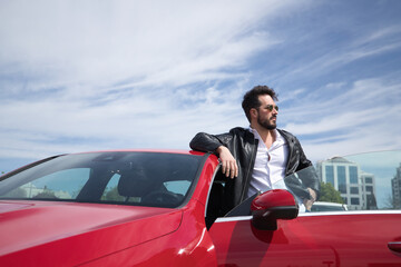 Handsome young man with beard, sunglasses, leather jacket and white shirt, leaning on the roof of his red sports car. Concept beauty, fashion, trend, luxury, motor, sports.