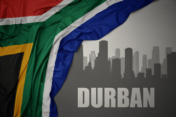 abstract silhouette of the city with text Durban near waving colorful national flag of south africa on a gray background. - obrazy, fototapety, plakaty