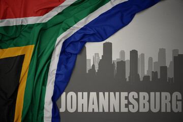 Obraz premium abstract silhouette of the city with text Johannesburg near waving colorful national flag of south africa on a gray background.