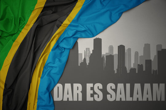 abstract silhouette of the city with text Dar es Salaam near waving colorful national flag of tanzania on a gray background.