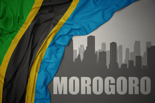 abstract silhouette of the city with text Morogoro near waving colorful national flag of tanzania on a gray background.