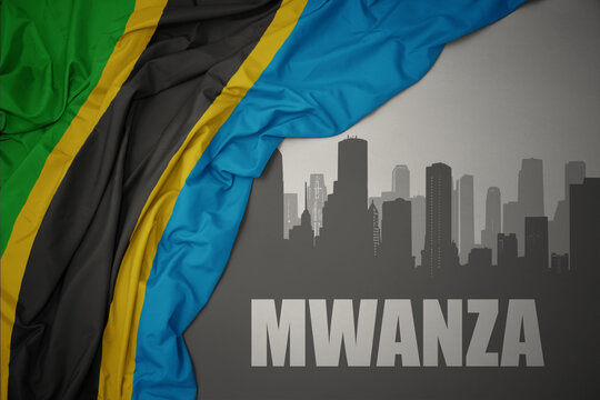 abstract silhouette of the city with text Mwanza near waving colorful national flag of tanzania on a gray background.
