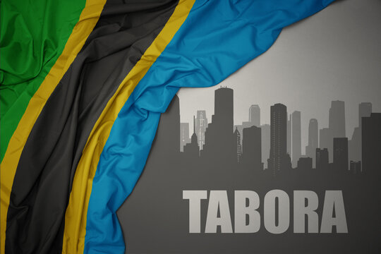 abstract silhouette of the city with text Tabora near waving colorful national flag of tanzania on a gray background.