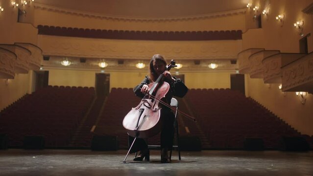 lonely woman is playing violoncello in empty music hall of opera house, female cellist is rehearsing