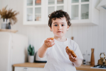 Little curly caucasian boy in white t-shirt at kitchen holding cookie in hand, offering a piece. Cute Italian kid, preschooler having breakfast at home, looking at camera, awakening. Childhood concept