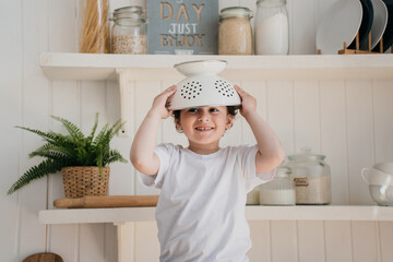 Little curly caucasian boy putting colander on his head at kitchen. Cheerful preschooler naughty...