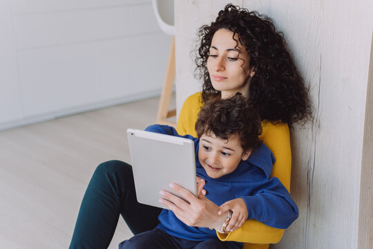 Young curly Spanish woman in yellow sweater and green pants sitting on floor at the kitchen with little son, showing him cartoons. Student  learning at home, take care a boy in blue looking at tablet.