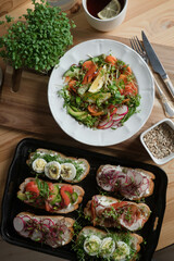 sandwiches with salmon, avocado, radishes, tomatoes, feta cheese, bacon, microgreens and eggs on a black tray. Fresh salad with seeds and lemon. Beautiful table setting. Top view.