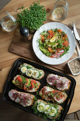 Open sandwiches with salmon, avocado, radishes, tomatoes, feta cheese, bacon, microgreens and eggs on a black tray. Fresh salad with seeds and lemon. A variety of healthy food.