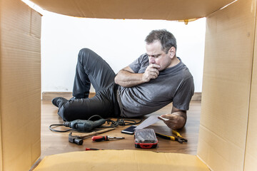 The man is lying on the floor with the tools around and thoughtfully studying the instructions,...