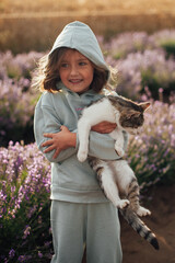 cute little girl in mint hoodie and pants with a cat in hands is standing among lavender field in morning light