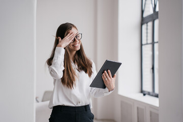 Grateful  young businesswoman eyes closed touching her head in excited expression talking by tablet making video call, received great news. Satisfied  girl in white shirt, black pants remote studying.