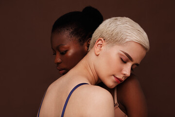 Two young women with different skin color standing together. Females put their heads on each other's shoulders with closed eyes.