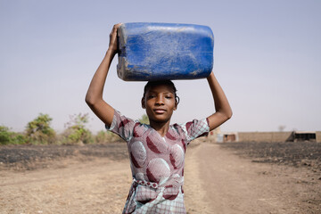 Young African girl carrying a heavy water container on her head, symbolising traditional roles of...